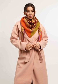 Cappotto di lana rosa con cintura in vita, impreziosito da un grande sciarpone multicolore con motivi in toni di arancione, verde e viola, appoggiato intorno al collo.