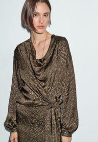 Donna con capelli corti castani che indossa un abito avvolgente a maniche lunghe, di colore marrone con un motivo, con un nodo laterale, in piedi su uno sfondo leggero uniforme.