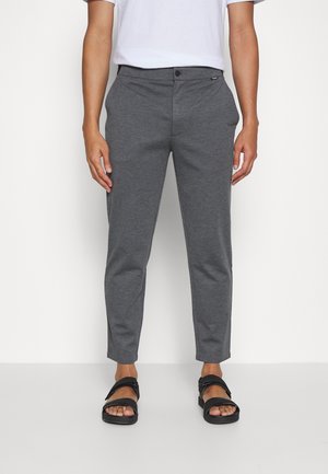 Homme portant un pantalon gris ajusté et des sandales noires à bouts ouverts, debout sur un fond blanc uni.
