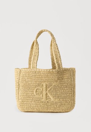 Sac cabas en paille tissée, couleur beige clair, doté de deux poignées et d'un logo "CK" en relief à l'avant. Surface texturée, forme rectangulaire.