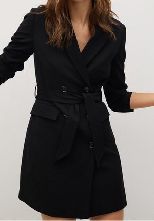 Kobieta w czarnej, dwurzędowej sukience-żakiecie z paskiem w talii i rękawami o długości trzy czwarte na jednolitym tle.