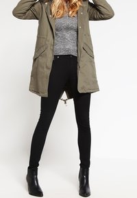 Olivgrön parka med huva, blixtlås fram och sidofickor, i kombination med tighta svarta jeans och svarta ankelstövlar.