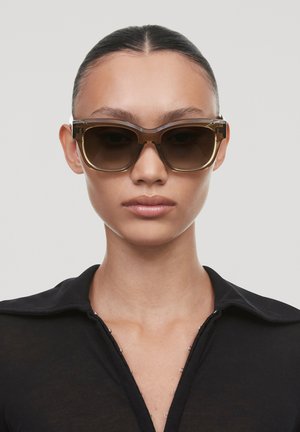 Bruine oversized zonnebril met een dikke, transparante montuur. Donker getinte lenzen. Model draagt een zwart col-shirt met knoopaccenten.