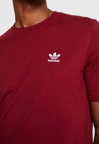 Camiseta de algodón burdeos con cuello redondo y mangas cortas, que presenta un logo trefoil blanco de Adidas en el lado izquierdo del pecho.