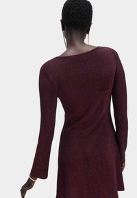 Burgundowa sukienka z długim rękawem o luźnym kroju, wykonana z połyskującego materiału. Posiada okrągły dekolt i asymetryczny dół.