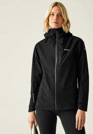 Mujer con una chaqueta negra para exteriores sosteniendo una botella de agua plateada contra un fondo claro y liso.