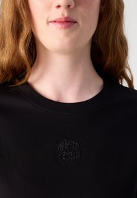 Svart t-skjorte med rund hals. Har en teksturert svart logo på brystet. Stoffet virker mykt og jevnt med en lett glans.
