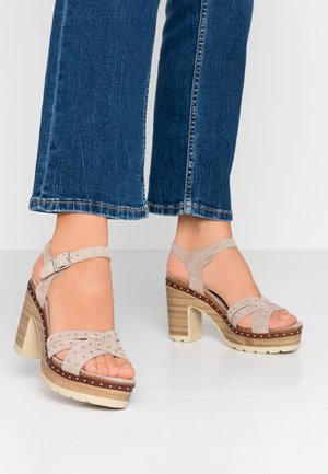 Fødder iført beige nitterede plateau-sandaletter med hæl og ankelremme samt blå denimjeans, stående på et hvidt gulv.