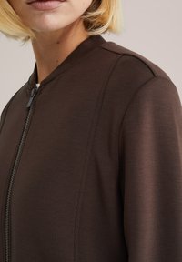 Rjava zadrga jakna z mehko teksturo, rebrastim ovratnikom in šivnimi detajli ob ramenih, s tesno prilegajočim se krojem.