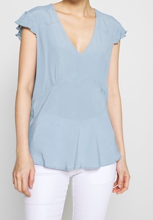 Blouse bleu clair à manches courtes évasées et encolure en V, portée avec un pantalon blanc, visible du cou aux hanches sur un fond uni.