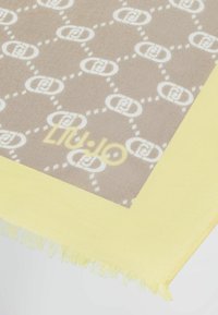 Bufanda marrón con patrón y diseño de logo blanco, con un borde amarillento con flecos y textura ligera. Nombre de la marca impreso en amarillo.