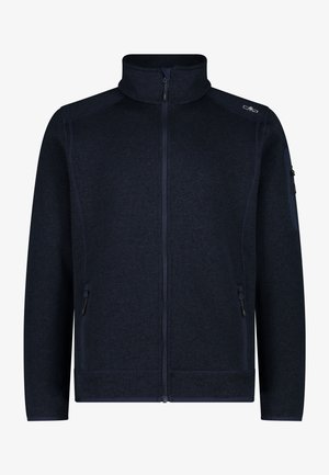 Hand in die Reißverschlusstasche einer dunkel marineblauen, strukturierten Fleecejacke gesteckt, getragen über schwarzen Hosen.