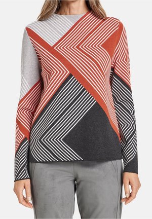 Kvinde iført en langærmet top med et abstrakt geometrisk mønster i rust, sort, grå og hvid, kombineret med grå bukser.