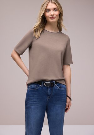 Blondynka w taupe krótkim rękawem koszuli wpuszczonej w niebieskie dżinsy z czarnym paskiem, stojąca na neutralnym tle.