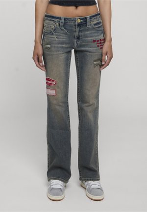 Femme portant un jean bleu délavé avec des patches rouges et des effets d'usure, associé à des baskets grises et blanches sur un fond clair uni.