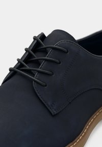Gros plan d'une chaussure habillée en cuir noir avec des lacets noirs et des coutures apparentes sur le côté, avec un bord de semelle marron.