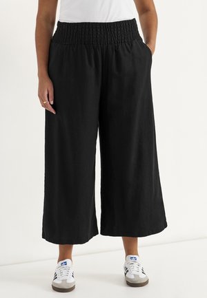 Osoba nosząca czarne, szerokie, krótkie spodnie z elastycznym paskiem, białe trampki z niebieskim logo i biały top, ręce w kieszeniach.