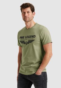 Zielony T-shirt bawełniany z czarnym nadrukiem tekstu "PME LEGEND AMERICAN CLASSIC" oraz grafiką skrzydła. Krótkie rękawy, okrągły dekolt, luźny krój.