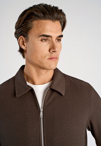 Bruine jas met een ritssluiting aan de voorkant, een kraag en een gestructureerde stof. Draagt over een wit shirt met een minimalistische snit.