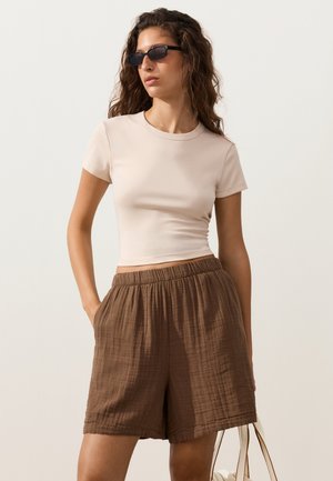 Femme portant des lunettes de soleil noires, un top beige ajusté à manches courtes et un short marron ample, debout les mains dans la poche devant un fond uni.
