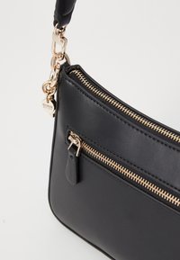 Svart lær crossbody-veske med gullfarget glidelås, avtagbar stropp og en liten anheng, med en glatt tekstur og elegant design.