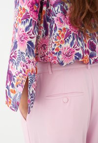 Blouse fleurie avec des motifs violets, roses et orange, associée à un pantalon rose pâle avec une poche arrière et un détail de bouton.