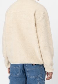 Giacca di pile crema con colletto alto, vestibilità rilassata, superficie texturizzata e cucitura sulla spalla, abbinata a pantaloni in denim.