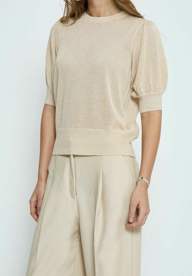 Maglione beige a maniche corte con scollo tondo, caratterizzato da una maglia strutturata e maniche leggermente gonfie. Abbinato a pantaloni beige a gamba larga.