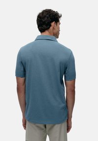 Rövid ujjú polo póló tompa kék színben, klasszikus gallérral, sima textúrával és laza fit-tel, hátulról bemutatva.