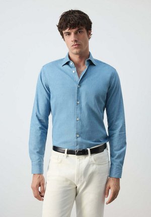 Giovane uomo che indossa una camicia azzurra con bottoni e pantaloni bianchi con cintura nera, in piedi davanti a uno sfondo chiaro uniforme.