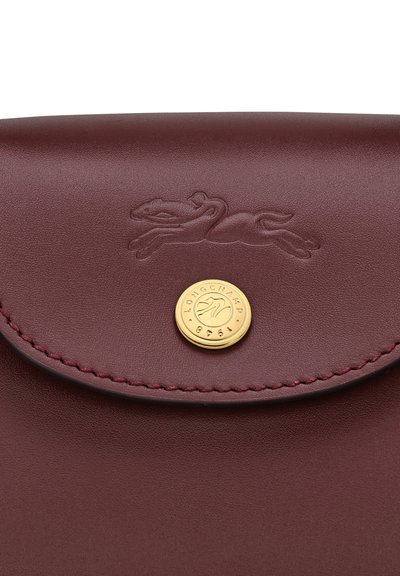 Burgundy leather bag with embossed logo, circular gold snap closure, and contrasting stitching along the edge. Smooth texture.