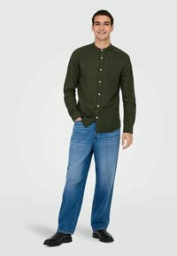 Giovane uomo in piedi, con una camicia verde scuro a bottoni, jeans blu e scarpe nere, sorride con una mano in tasca.