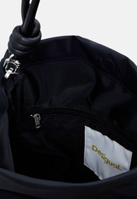 Interno di una borsa di stoffa nera con una tasca con zip e un'etichetta bianca visibile con la scritta "Desigual." Texture liscia, design minimalista.