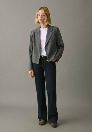 Blazer grigio accorciato con due bottoni, abbinato a una camicia bianca e jeans a zampa d'elefante scuri. Il modello indossa scarpe marroni con borchie decorative.