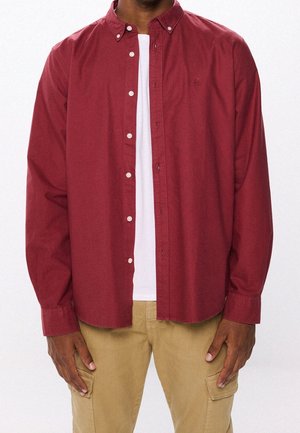 Homme portant une chemise rouge à manches longues déboutonnée par-dessus un t-shirt blanc, avec un pantalon cargo beige, debout devant un fond uni.