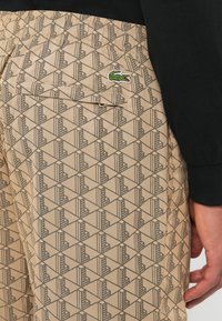 Pantaloni corti beige con motivo geometrico e piccolo logo di coccodrillo verde sulla tasca, indossati con una camicia nera a maniche lunghe.