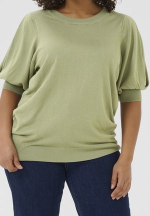 Person trägt einen hellgrünen Kurzarm-Pullover mit Puffärmeln und dunkelblaue Jeans, steht mit gelockerten Händen an den Seiten.
