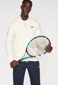 Ένα κρεμ μπουφάν με φερμουάρ και ψηλό γιακά, συνδυασμένο με σκούρο μπλε παντελόνι φόρμας με μικρό λογότυπο, και μια μαύρη ρακέτα τένις με μπλε λεπτομέρειες.