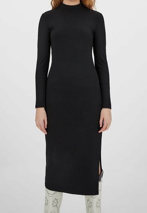 Kobieta w dopasowanej czarnej prążkowanej sukience midi z długim rękawem i bocznym rozcięciem, w zestawieniu z białymi butami we wzory.