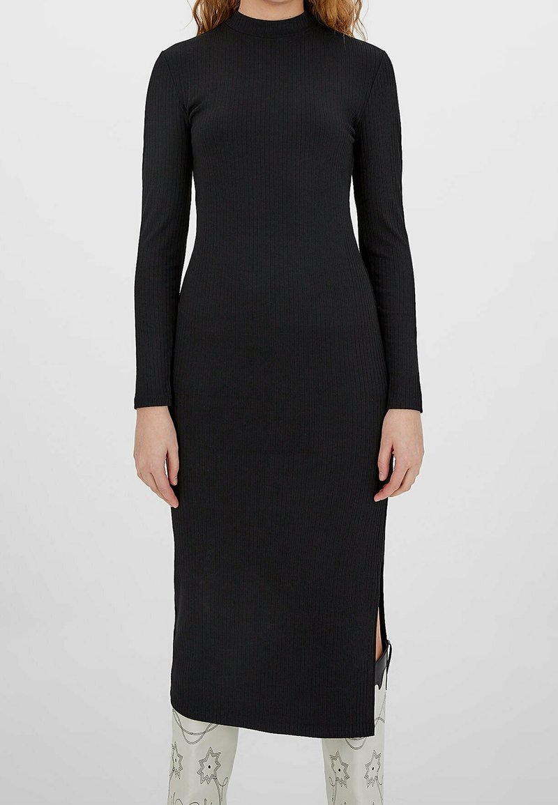 Femme portant une robe midi noire ajustée à manches longues côtelées avec une fente latérale, associée à des bottes blanches à motifs.