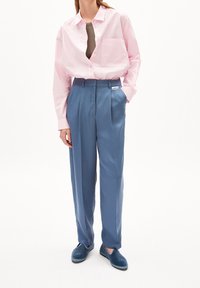 Žena v růžové košili na knoflíky, pod kterou má hnědý top, zasunutá do modrých kalhot s vysokým pasem, k nimž má ladící modré slip-on boty.