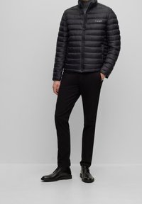 Veste matelassée noire avec coutures horizontales, col montant et fermeture éclair à l'avant, associée à un pantalon noir et des bottines, sur un fond gris.