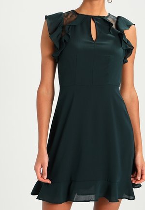 Femme portant une robe sans manches vert foncé avec des épaules à volants, des détails en dentelle près de l'encolure, et un ourlet évasé.