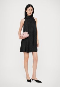 Vestido preto sem mangas com um colarinho alto, textura suave, silhueta evasê, combinado com uma pequena mala rosa e sapatos pretos de bico fino.