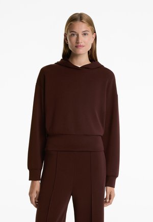 Rjava kapuca s kapuco, sproščenega kroja, z ribastimi manšetami in pasom, izdelana iz mehkega materiala; nosi se s sovpadajočimi hlačami.