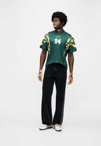 Πράσινη φανέλα με κίτρινες πινελιές και αριθμό 94· συνδυασμένη με σκούρο φαρδύ τζιν και λευκά αθλητικά παπούτσια. Ομαλή υφή υφάσματος, άνετη εφαρμογή.