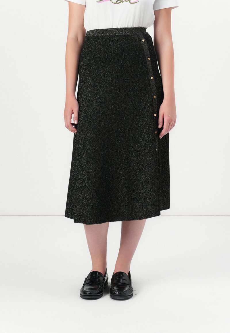 Una persona lleva puesta una camiseta gráfica blanca y una falda negra de longitud a la rodilla con botones dorados de un lado, combinada con unos mocasines negros.