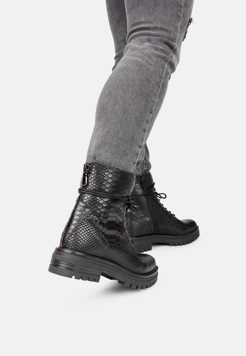 Bottines noires à lacets avec un motif en peau de serpent texturé, semelles en caoutchouc et détails en métal sur la fermeture éclair sur le côté, associées à un jean gris.
