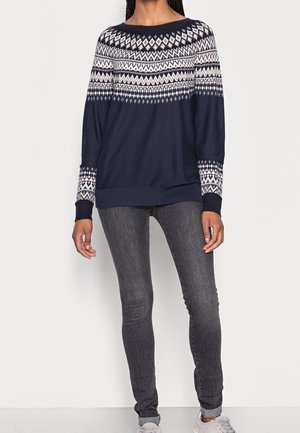Femme portant un pull bleu marine avec un motif géométrique blanc sur le haut de la poitrine et les manches, un jean skinny gris foncé et des baskets claires.