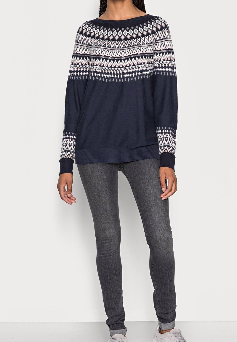 Femme portant un pull bleu marine avec un motif géométrique blanc sur le haut de la poitrine et les manches, un jean skinny gris foncé et des baskets claires.